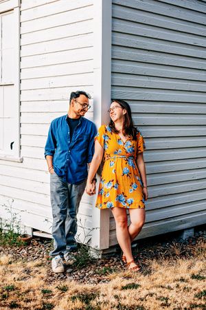 A couple wearing blue shirt and yellow floral dress pose casually against a white building wall outdoors.
