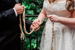Hands hold braided cord during handfasting unity ceremony at outdoor wedding with lace dress and formal attire.