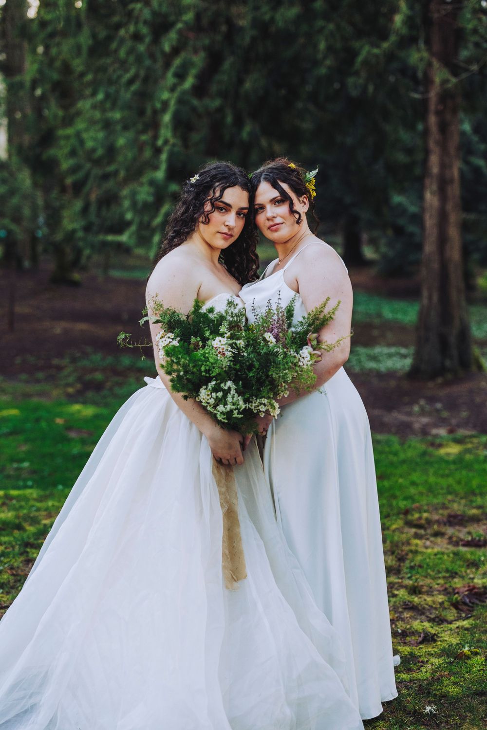 Two brides in flowing white wedding dresses pose together in a wooded outdoor setting while holding a green and white bouquet.