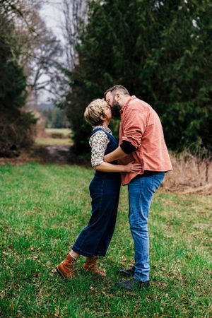 A romantic moment shared between two people embracing on grassy field with evergreen trees in background.