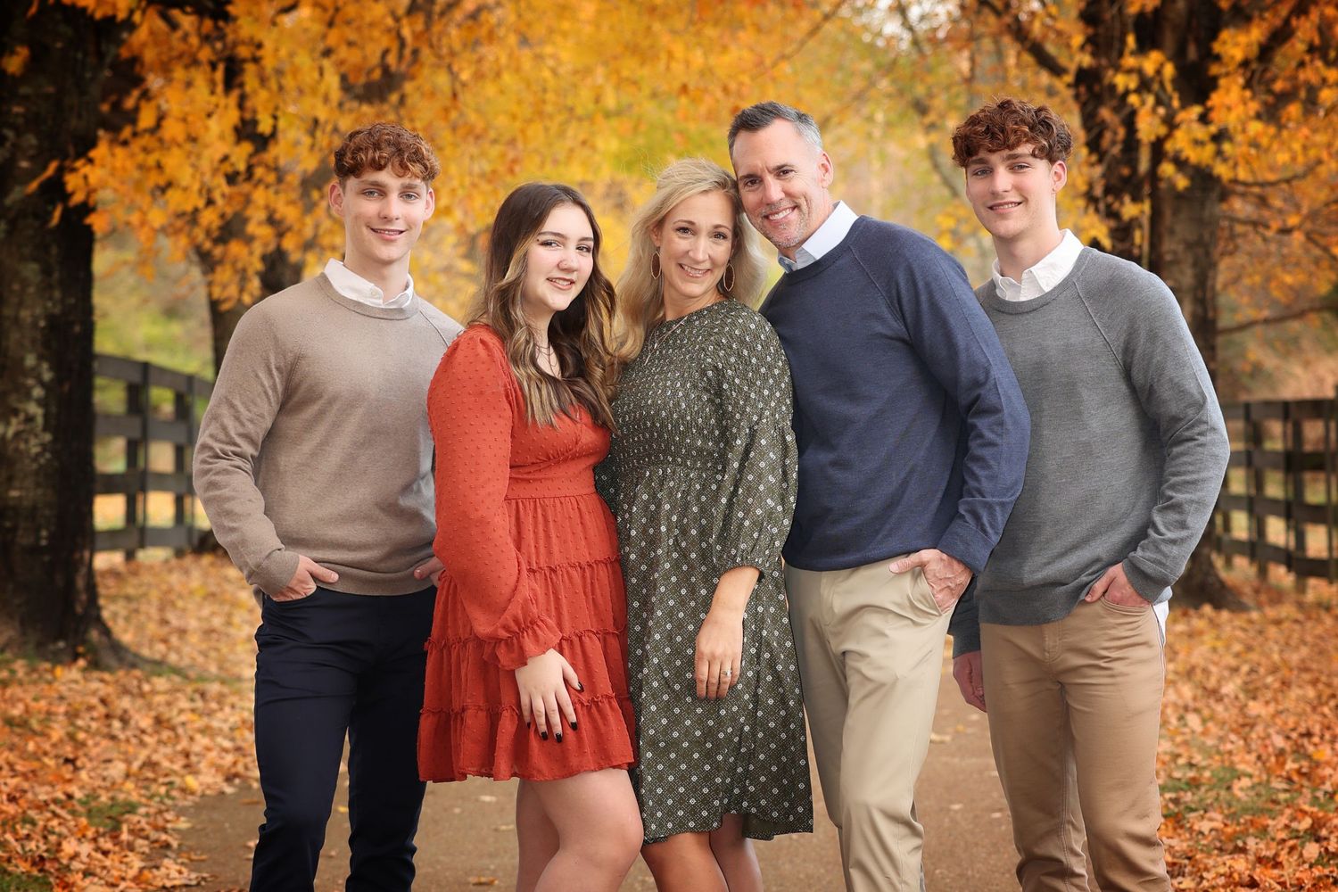 Family of five posing outdoors against autumn trees and a black fence in Fall Mini session photo shoot.