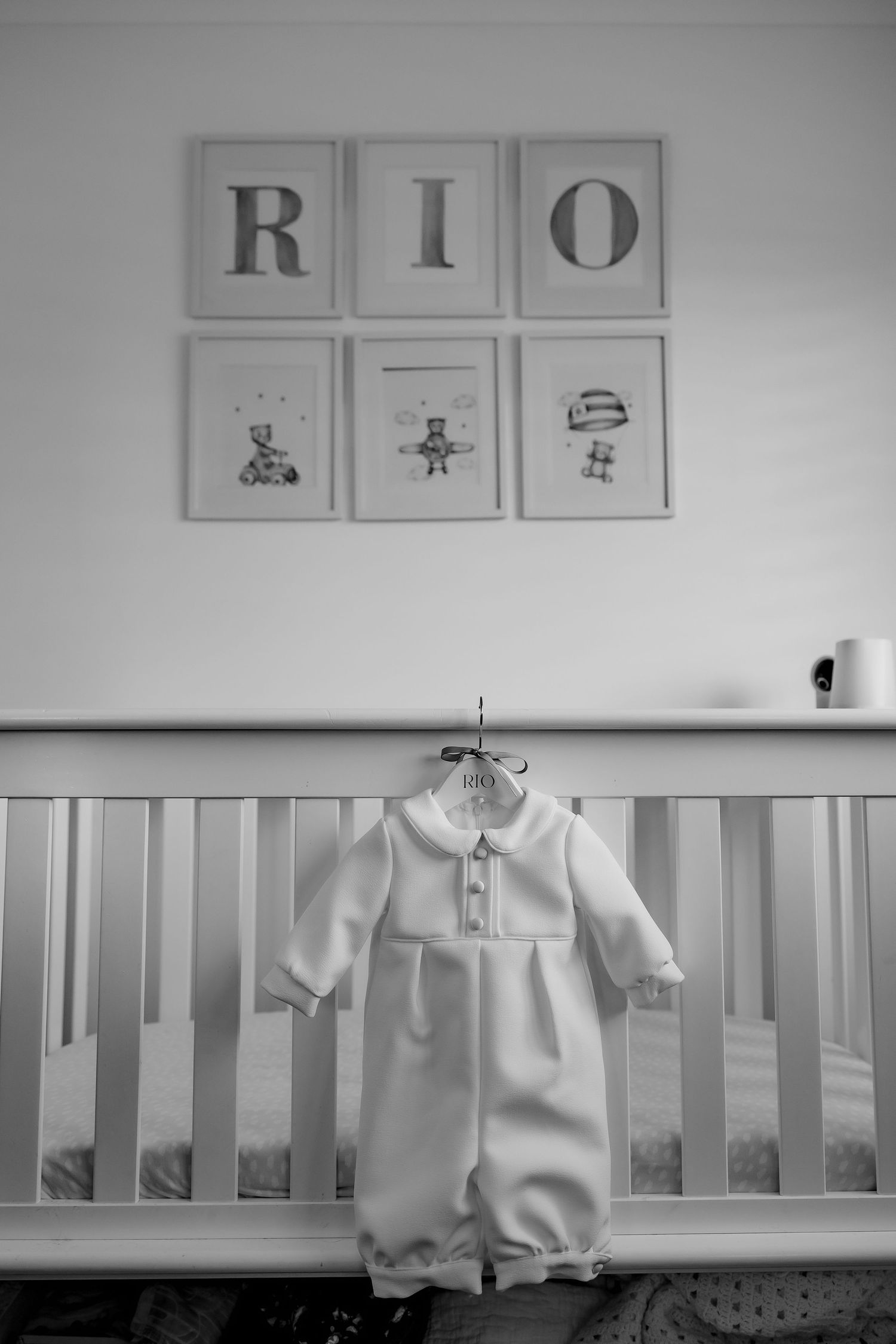 A black and white nursery photo showing 'RIO' wall art and a white baptismal outfit.