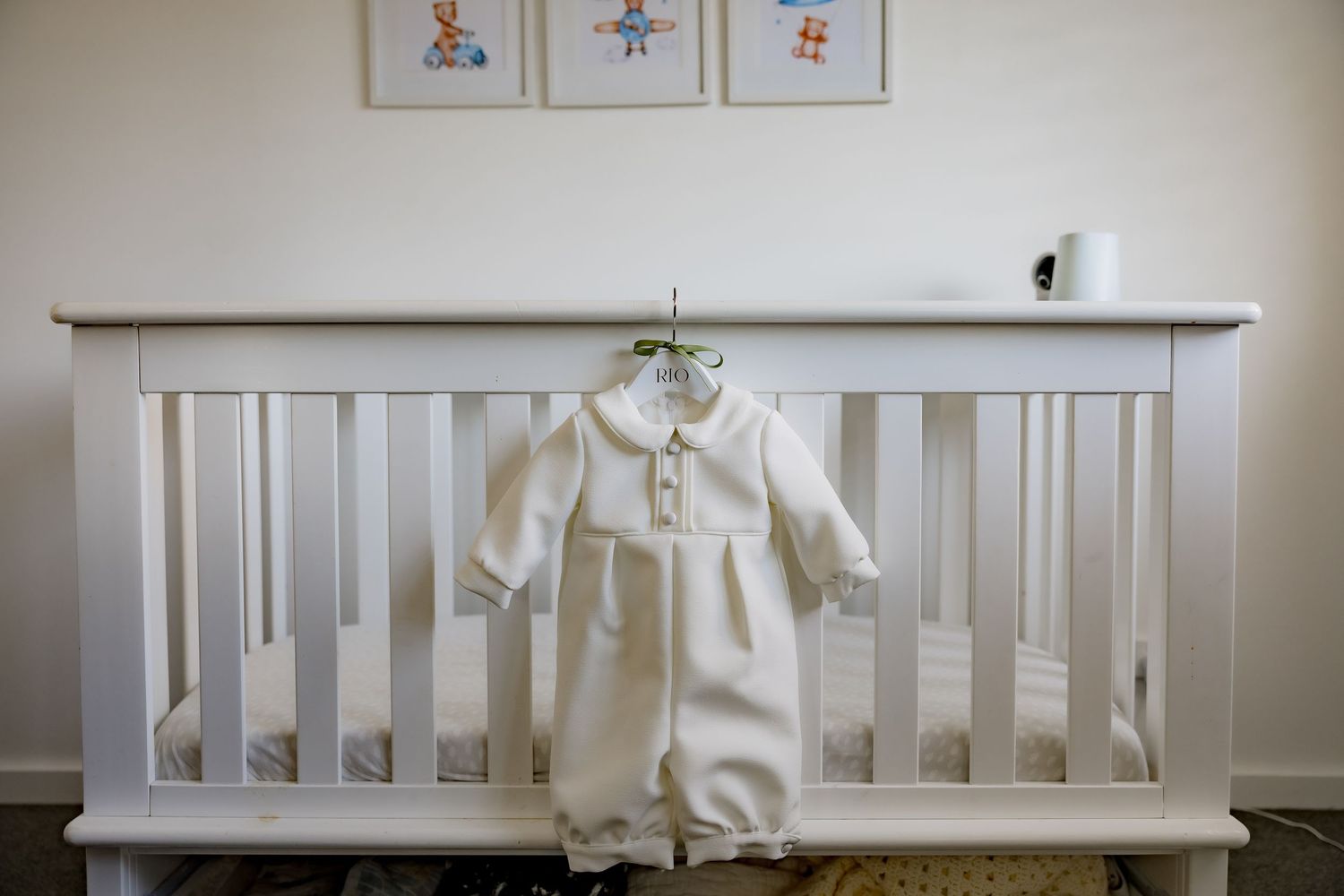 A white baptismal gown displayed on a white crib under framed nursery artwork.