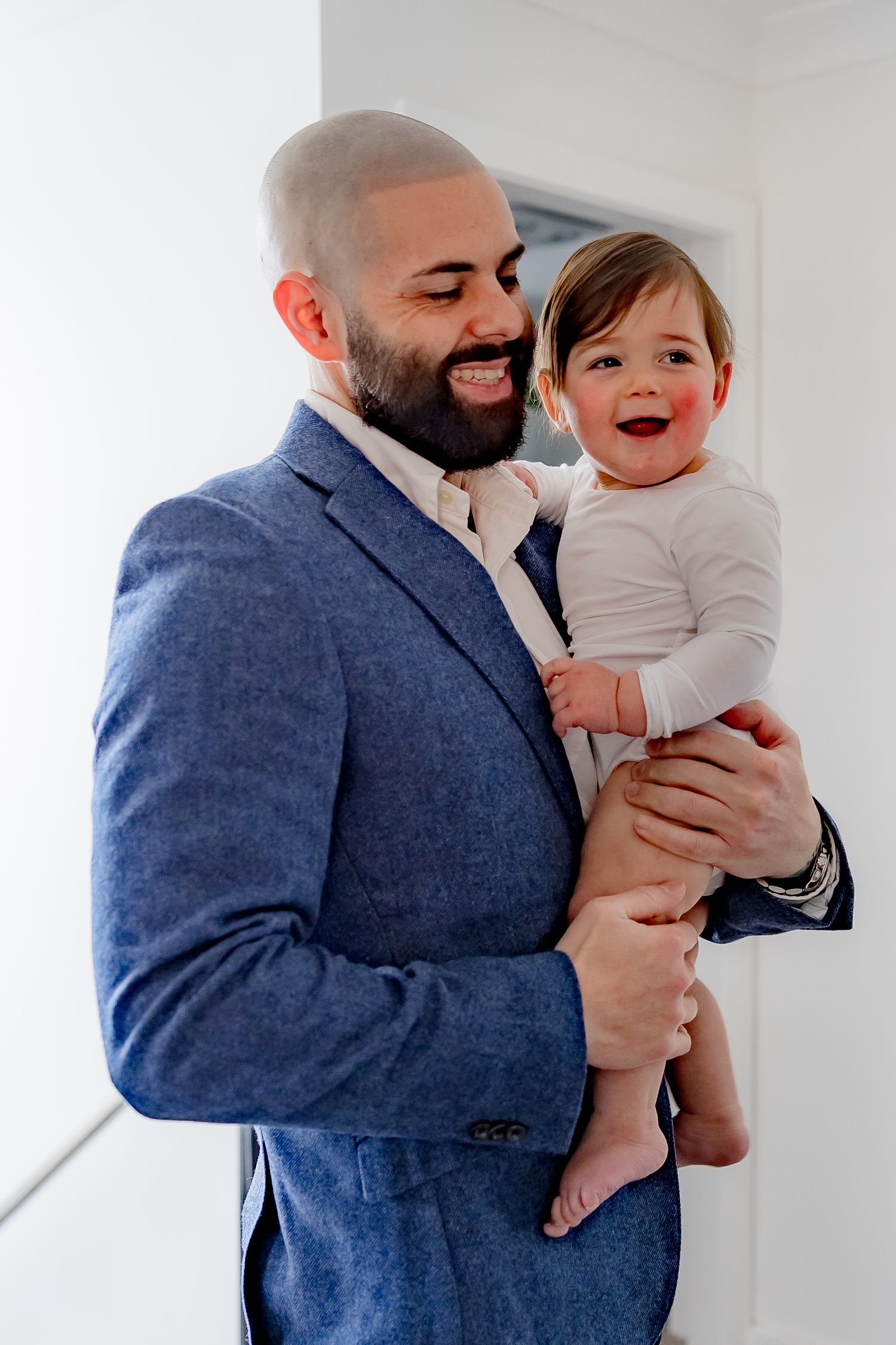 A joyful father in a blue blazer holds and smiles with his laughing baby wearing white.
