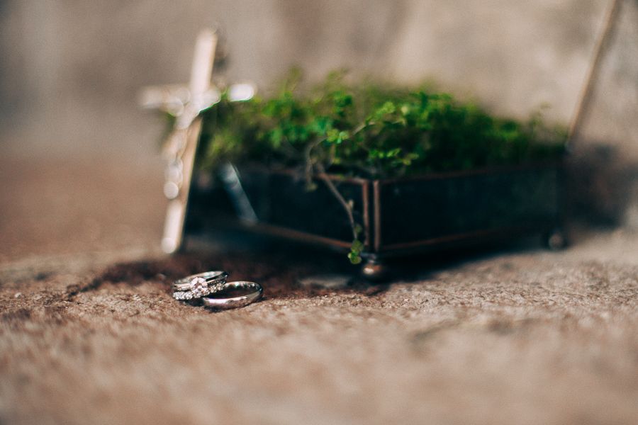 Wedding ring resting on sandy surface near small cross and green moss decor.
