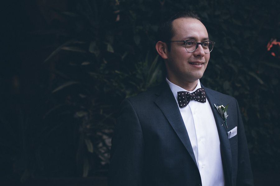 Dapper groom in black tuxedo with polka dot bow tie smiling outdoors near green foliage.
