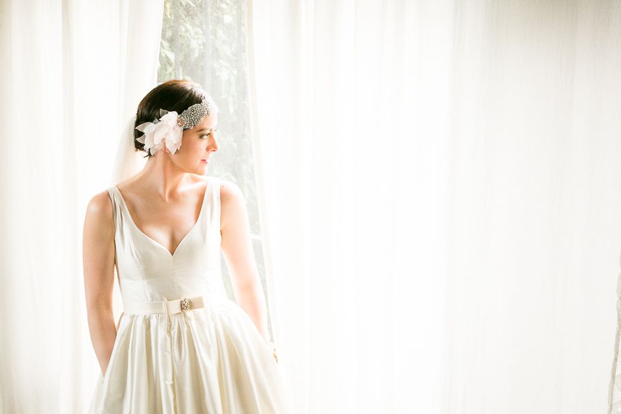Elegant bride in white gown with floral headpiece standing by bright window in soft light.