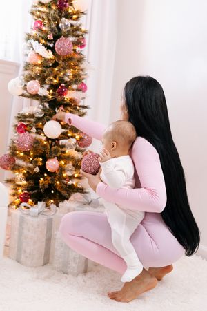 Parent and baby decorating pink and gold Christmas tree together in white room.