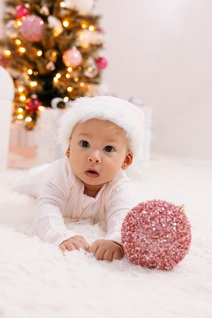 Baby in white outfit lying on fur rug with pink glitter ornament near Christmas tree.