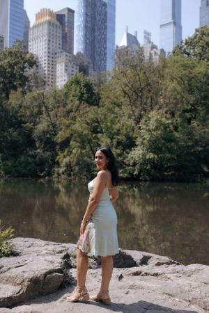 Person in a white dress poses on rocks by a lake in Central Park with NYC skyscrapers in the background.