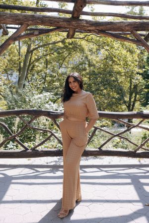 Person in a beige knit outfit poses under a rustic wooden pergola in a park setting.