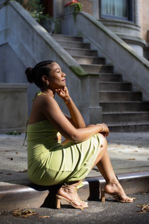 A woman in a lime green dress poses thoughtfully on steps in front of a brownstone building.
