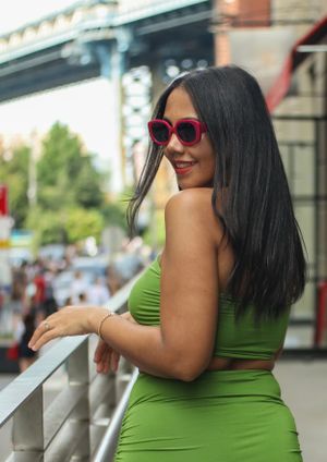 Woman in green two-piece outfit and red sunglasses poses by railing with urban backdrop.