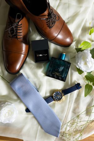 Groom's accessories including brown leather shoes, tie, watch and cologne on white fabric.