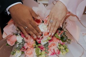 Close-up of hands with wedding rings resting on a pink and white floral bouquet.