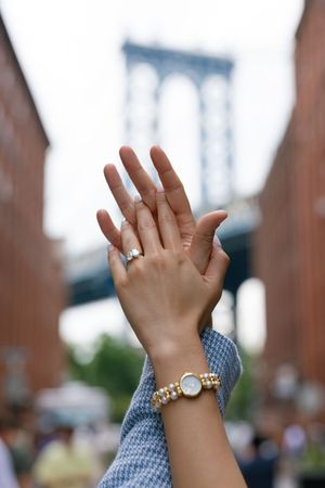Close-up photo showing hands wearing engagement ring and bracelet with Manhattan Bridge visible in background.