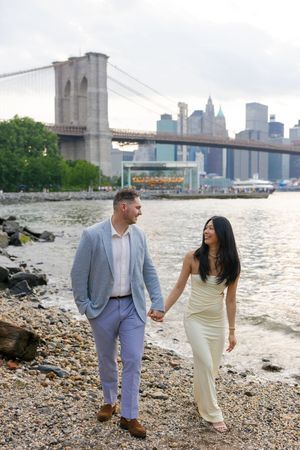 A couple shares a romantic moment along the Brooklyn waterfront with the Manhattan skyline and bridge in the background.