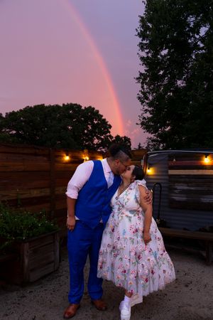 A couple shares a romantic moment under a pink sky with a vibrant rainbow and glowing lights in a rustic outdoor setting.