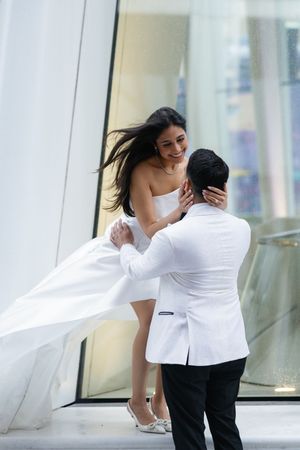 Playful newlywed couple embrace on a modern balcony as the bride's dress and hair flow in the wind.