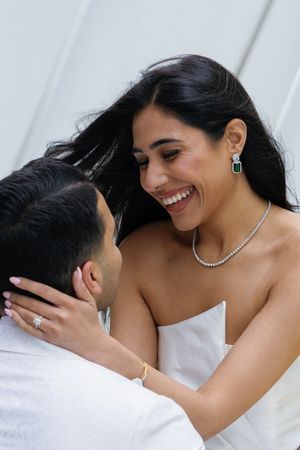 A joyful bride in white dress wearing emerald earrings and necklace shares an intimate moment with her partner.