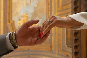 Hands delicately pass a small red object in an ornate gold-decorated room with luxurious architectural details.