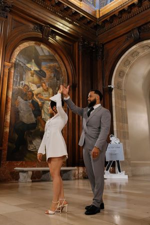 A couple dances together in an elegant ballroom with ornate walls and murals.