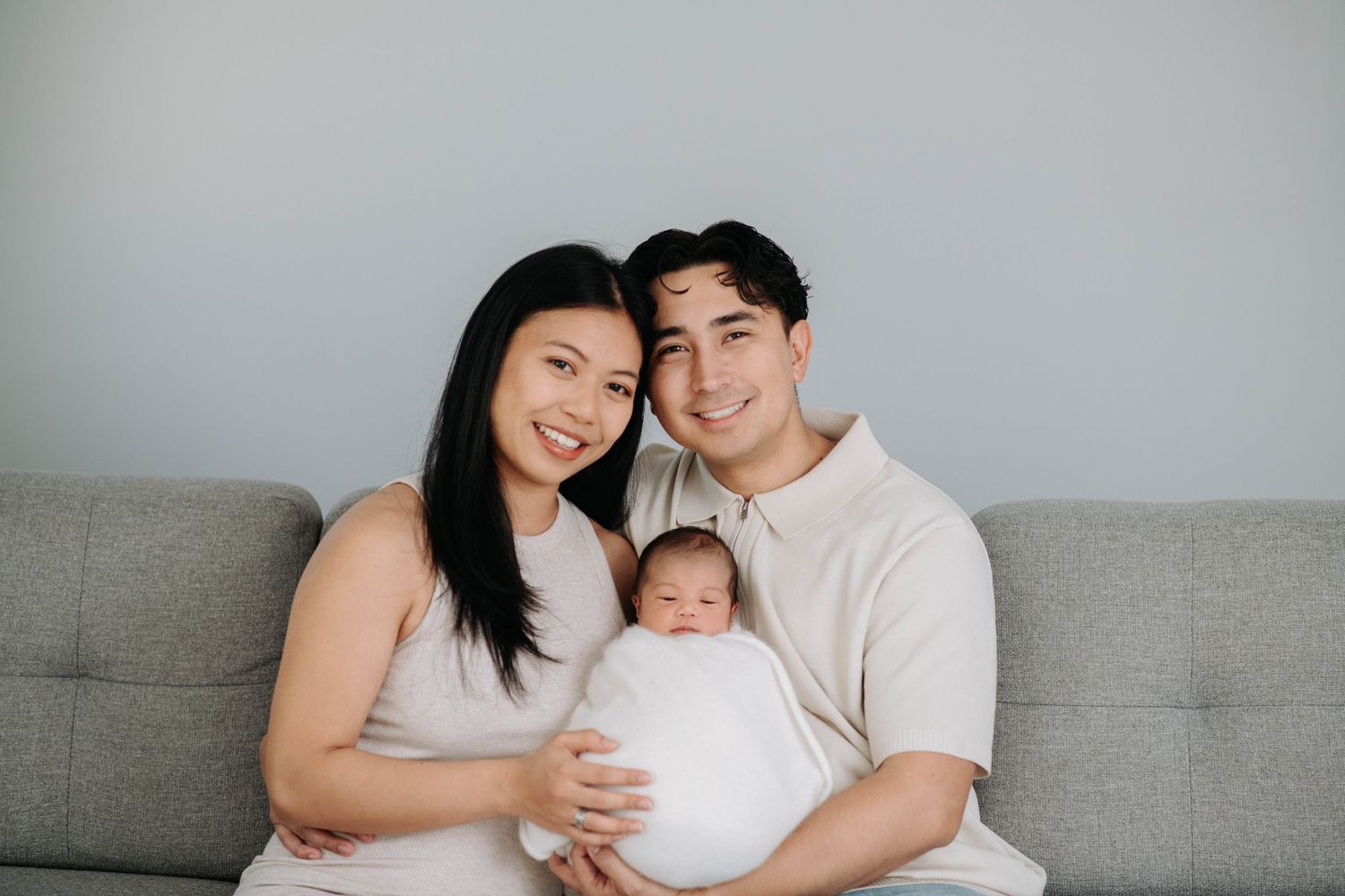A cozy family portrait with parents and newborn baby on grey sofa.