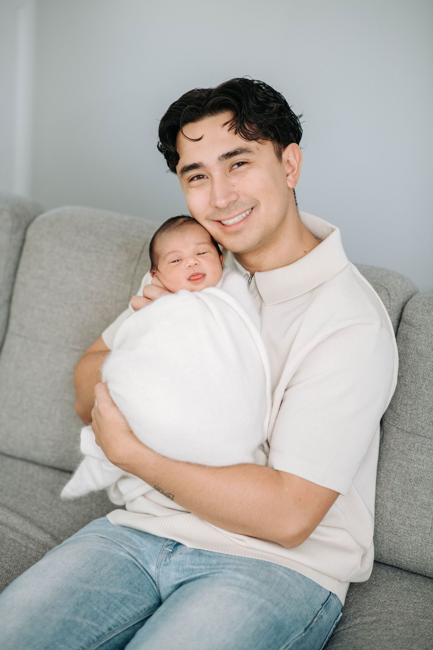 Parent sitting on grey sofa holding sleeping newborn in white swaddle.