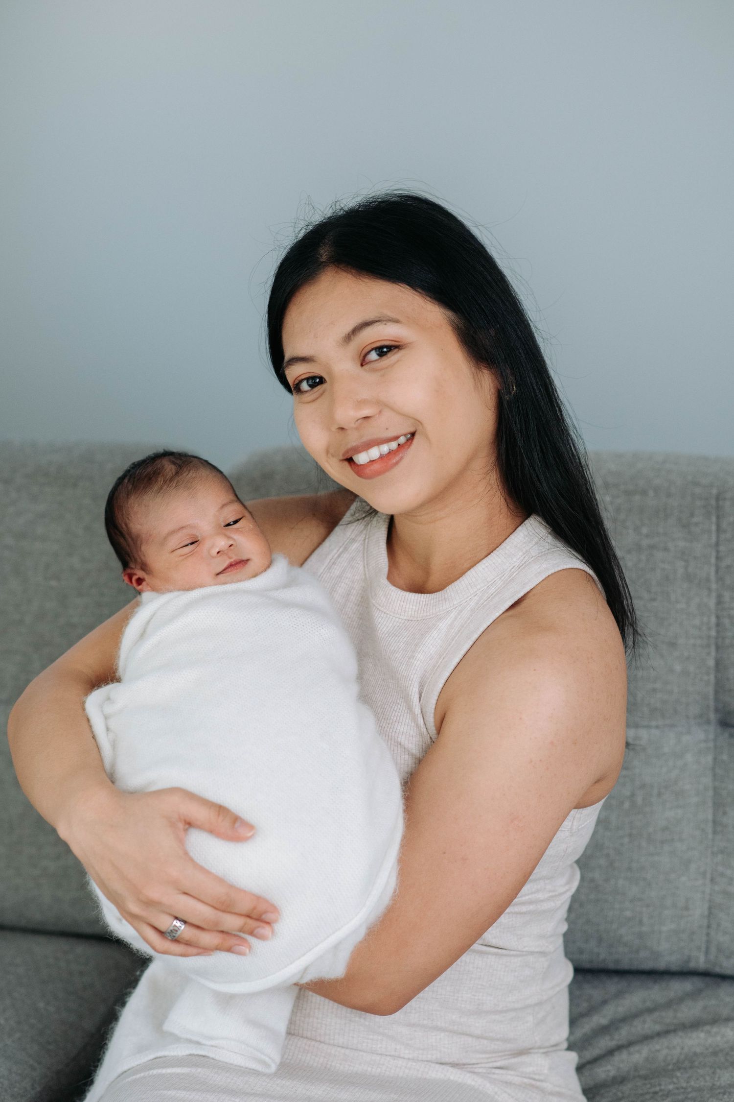 Mother holding newborn baby on grey sofa in minimalist setting.