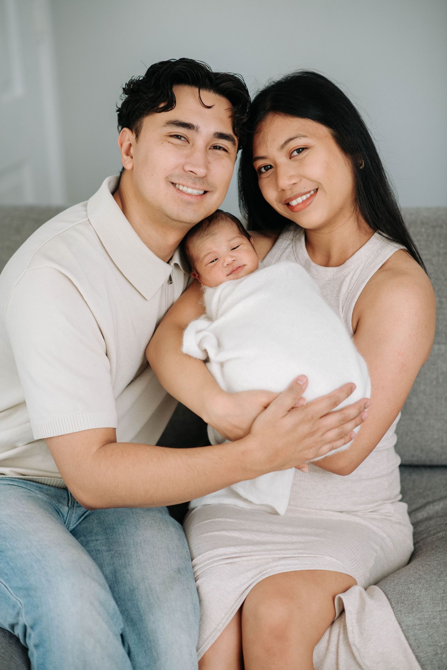 A family portrait showing loving parents with their newborn baby on a grey sofa.