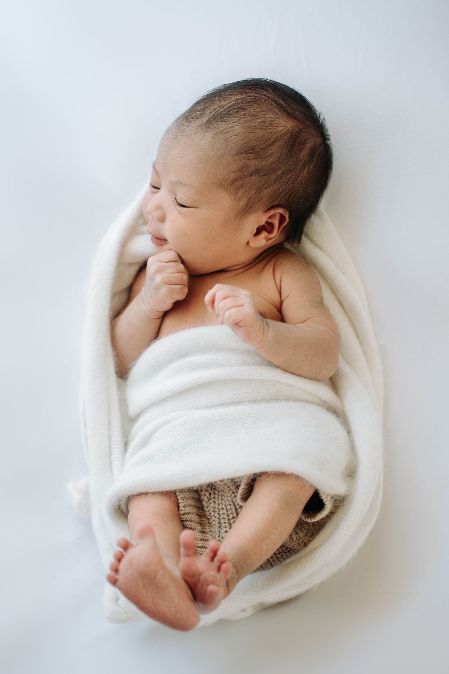 Sleeping newborn swaddled in white blanket with knit pants showing bare feet against white background.