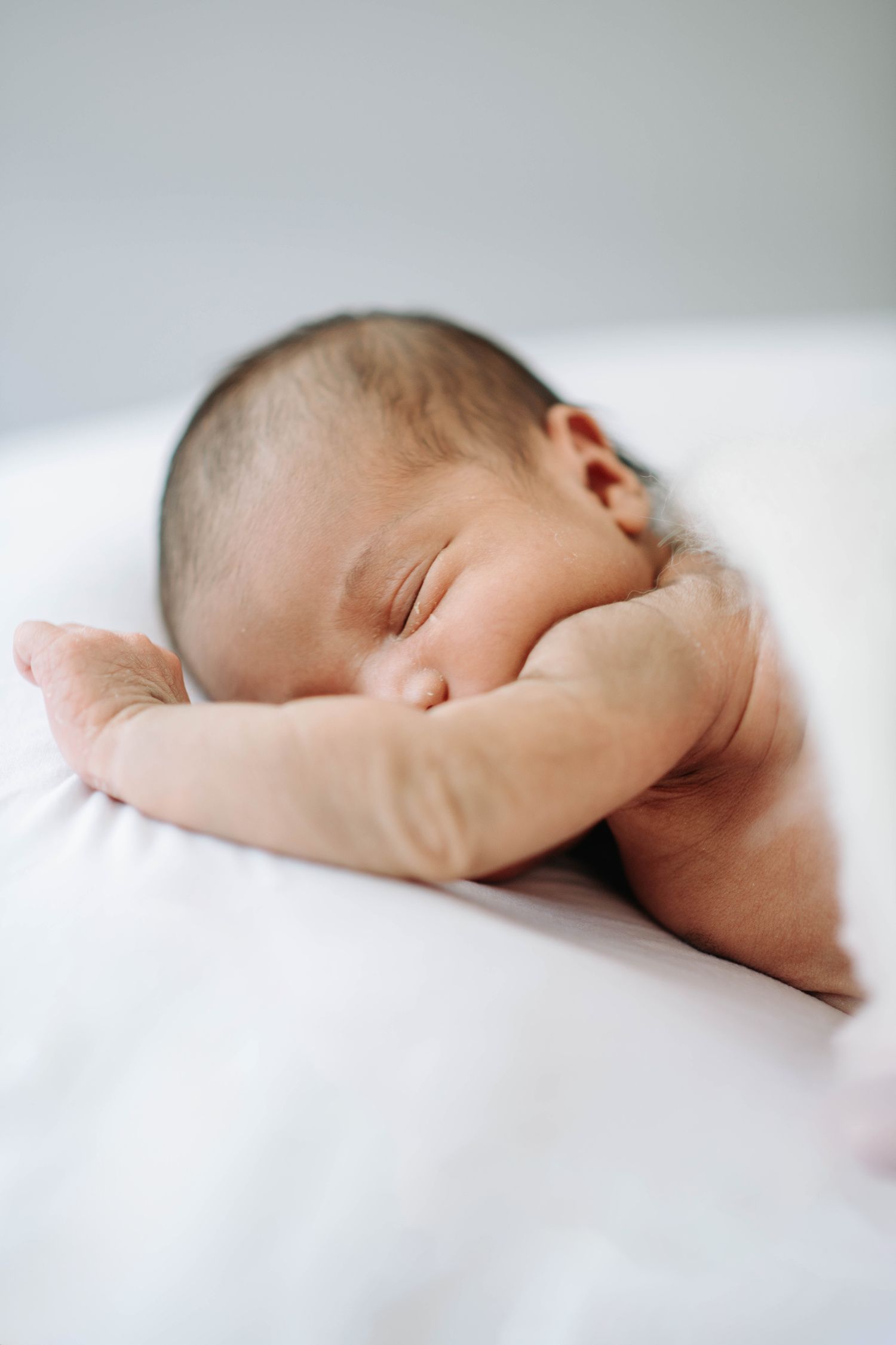 Sleeping newborn baby with arm raised resting on white bedding.