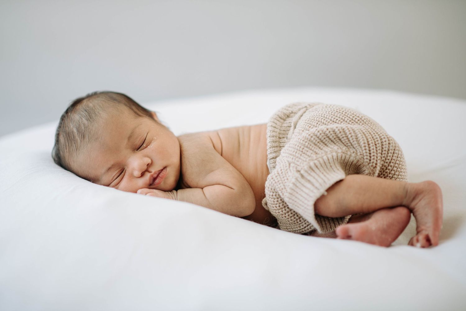 Peaceful newborn wearing knit shorts sleeping on white surface in natural light.