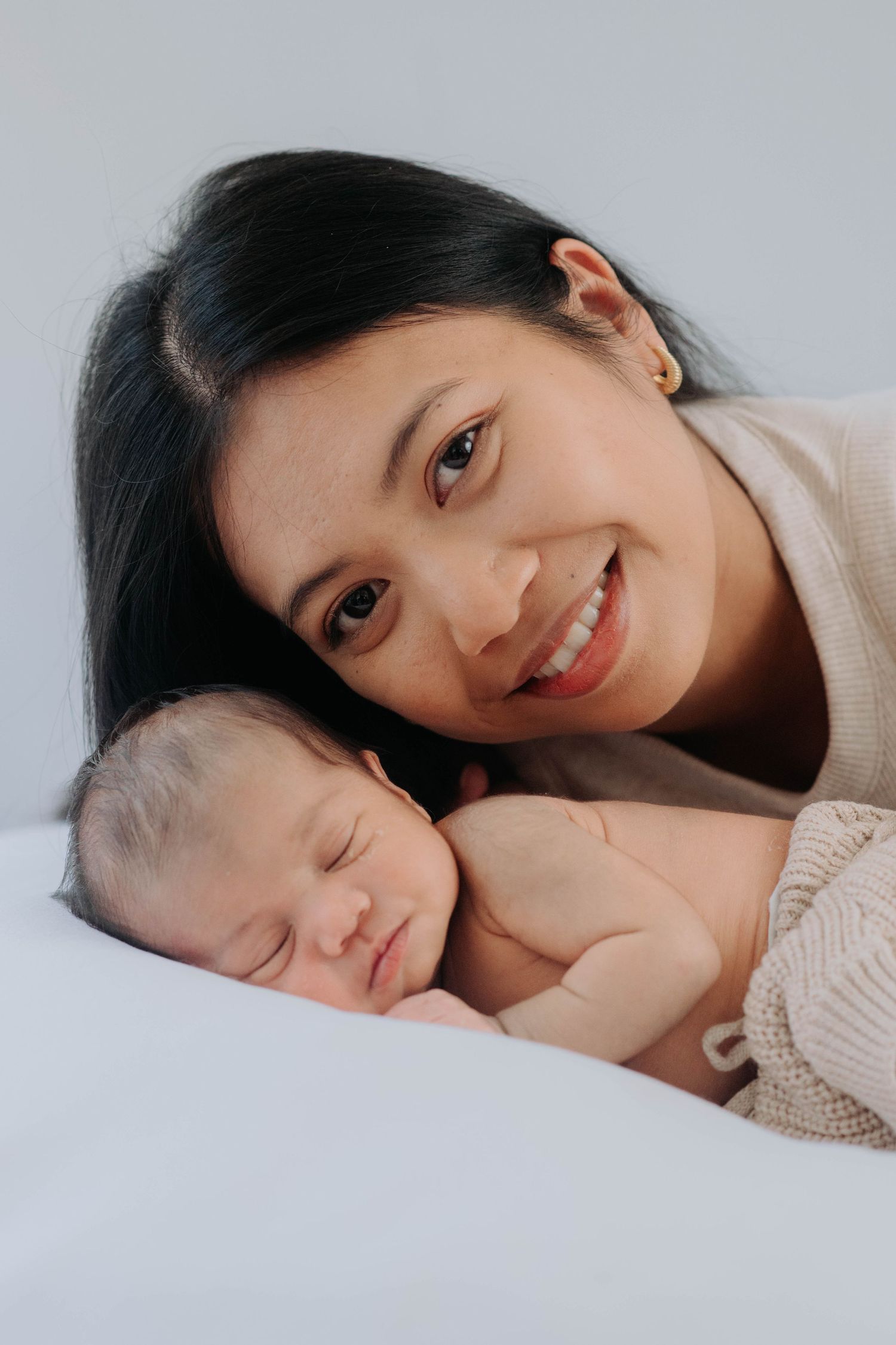 Mother and sleeping newborn share tender moment on white bedding.
