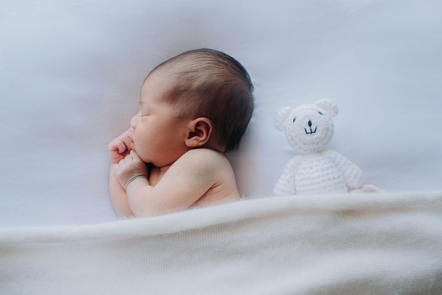 A peaceful newborn sleeping on soft white bedding next to a small white teddy bear.