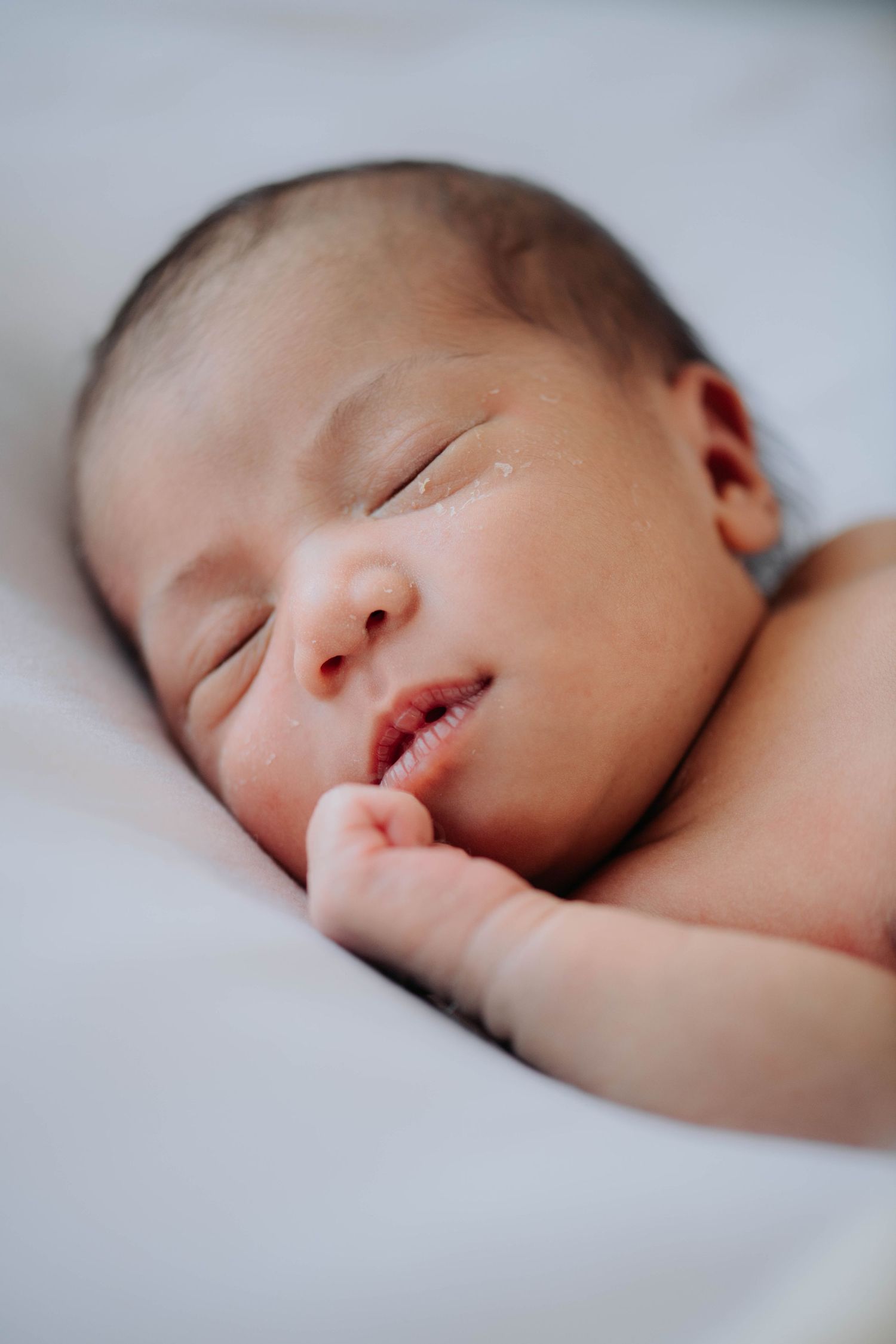 A sleeping newborn with hand raised to mouth on white bedding.