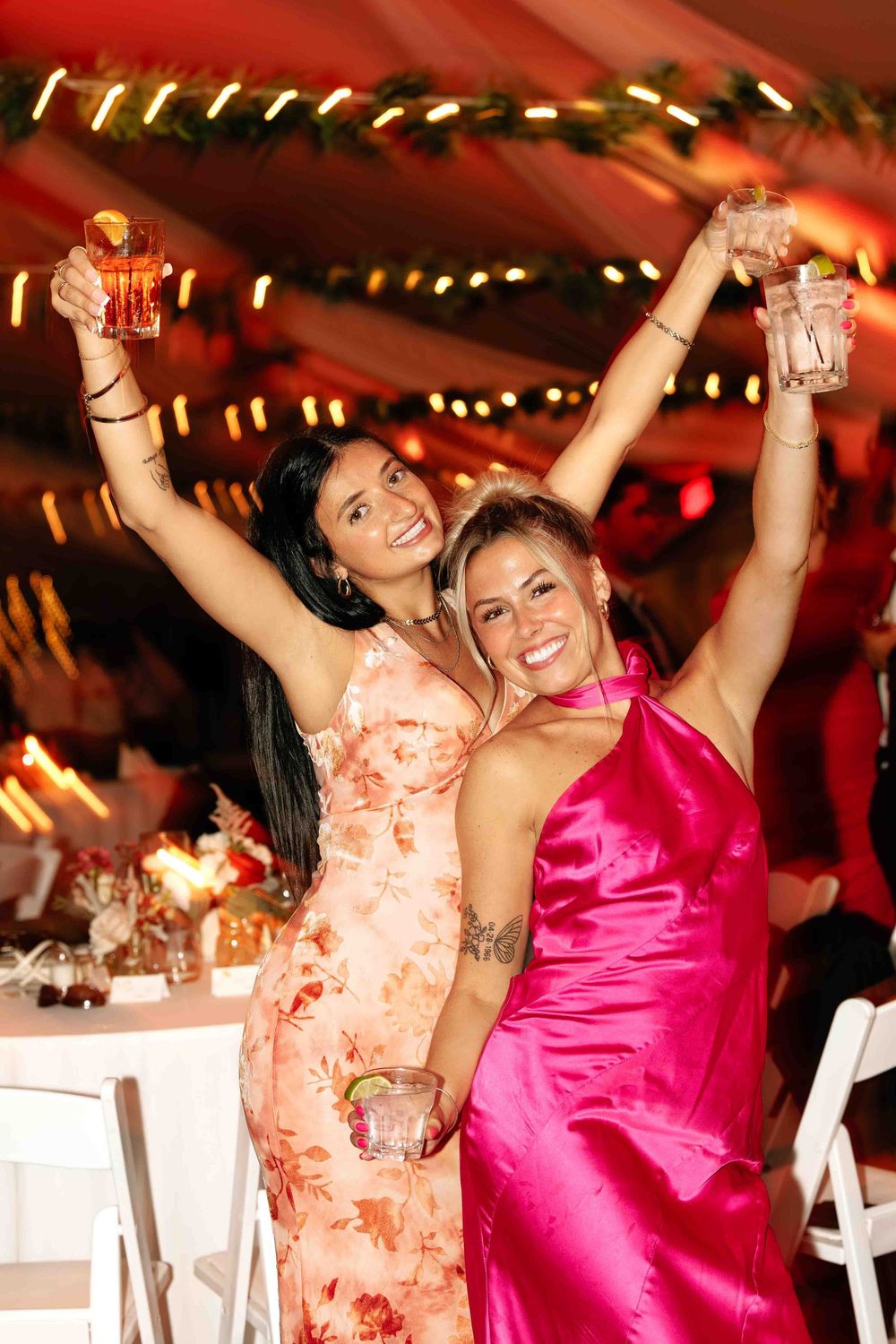 Two women in elegant dresses celebrate joyfully at an evening party with raised glasses under decorative lighting.