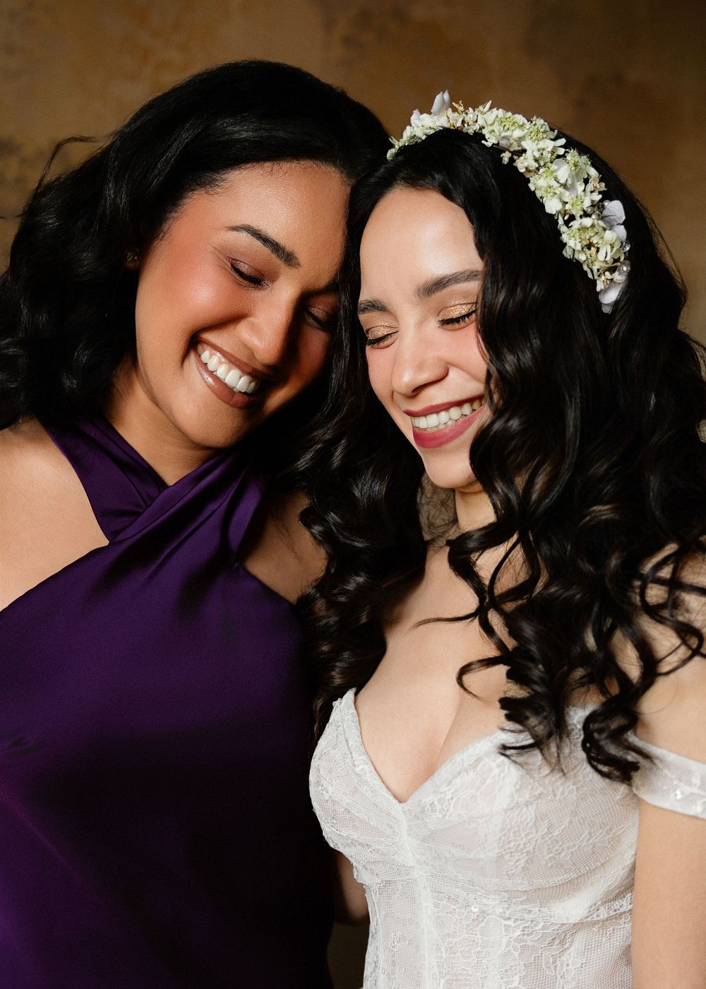 Two women with dark hair share a joyful moment, one in a white wedding dress with floral crown and one in purple.