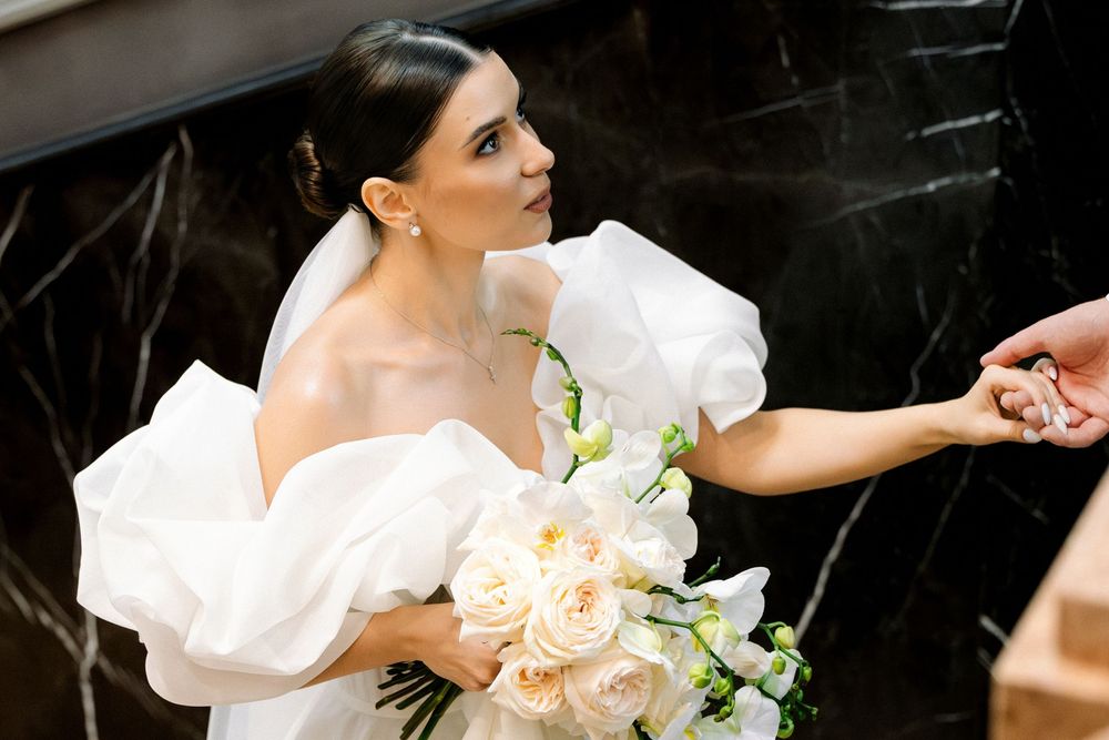 A bride in an elegant white off-shoulder gown holds a peach and white bouquet while reaching out her hand.