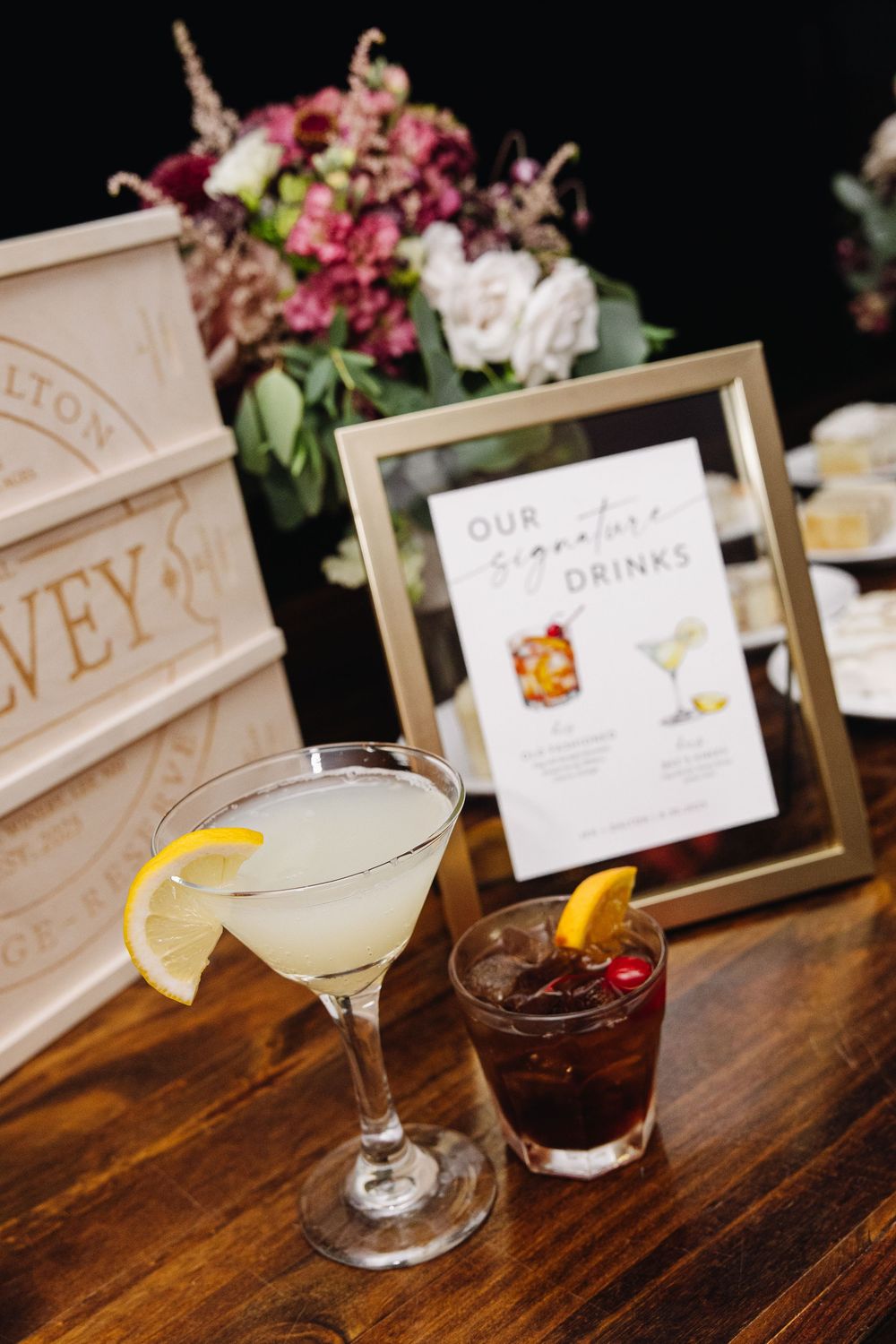 Elegant cocktails with garnishes displayed on a wooden bar top next to framed menu cards and floral arrangements.