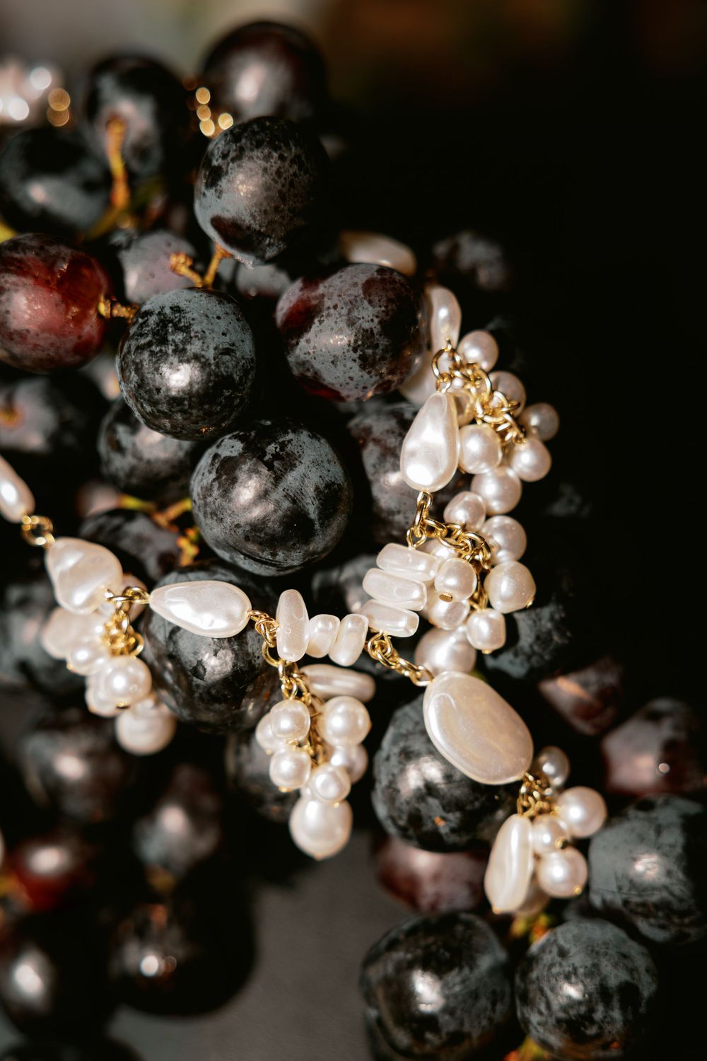 Pearl and gold jewelry displayed alongside dark purple grapes in moody lighting.