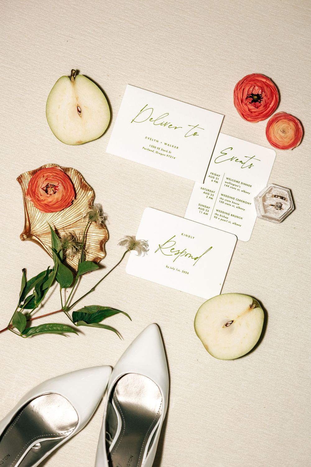 Flatlay of wedding stationery with red ranunculus flowers, sliced apples, and silverware on cream background.
