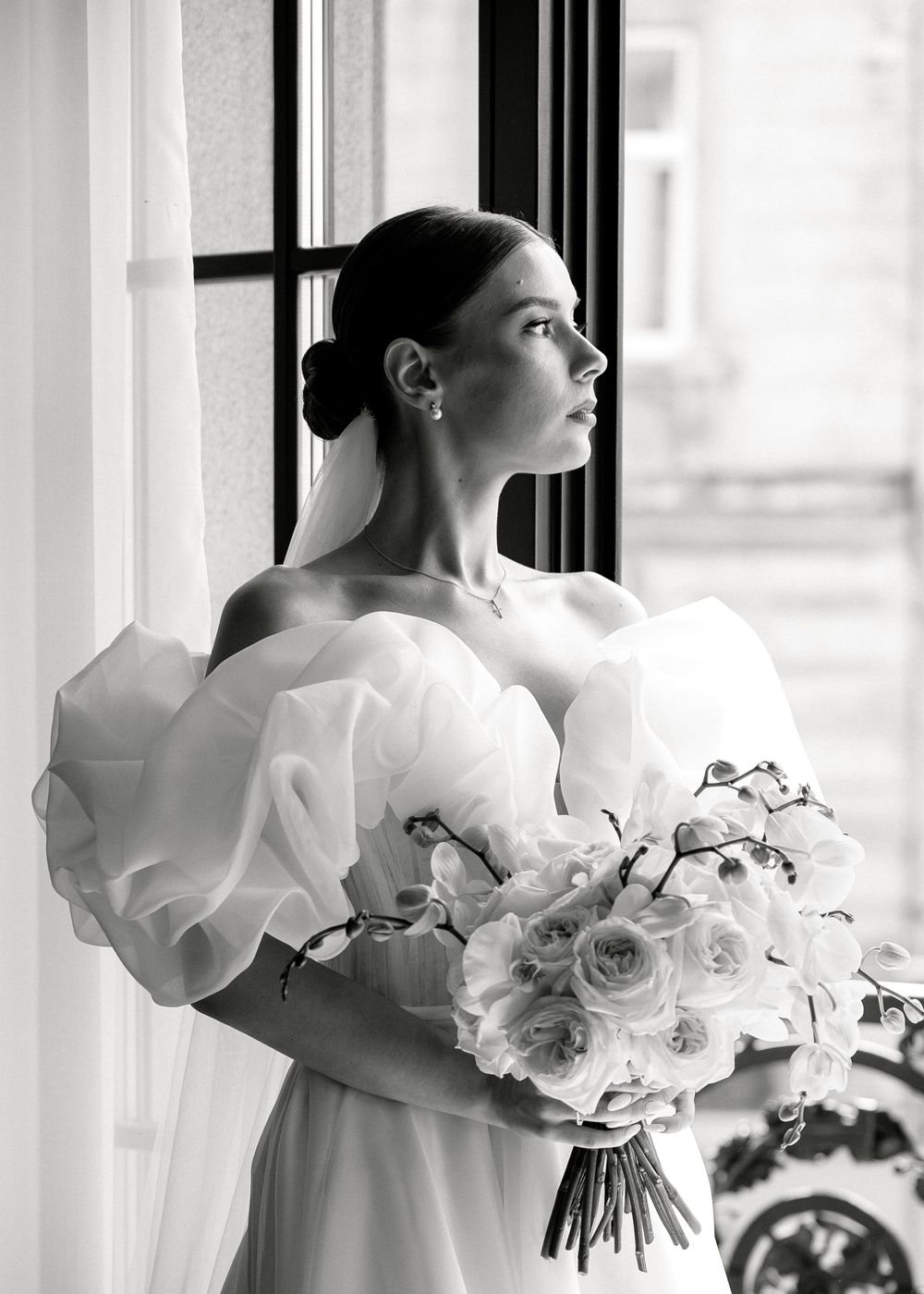 Elegant bridal portrait with white puffy sleeves and a bouquet of roses against sheer curtains in natural light.