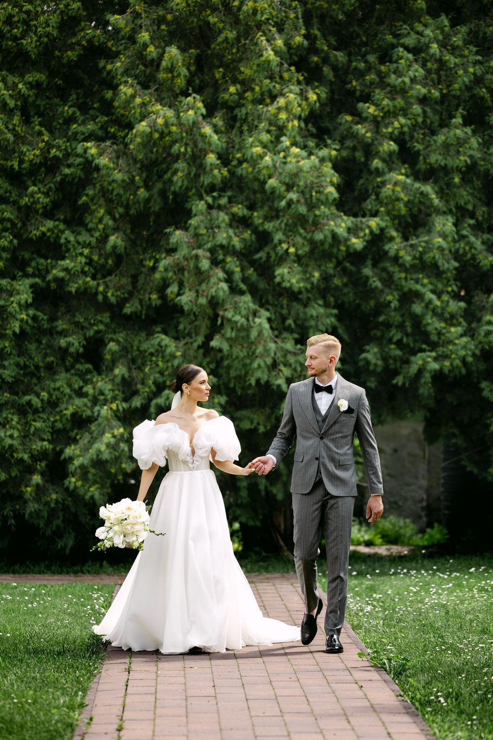 A newlywed couple walks hand in hand along a brick path surrounded by lush green trees and scattered flower petals.