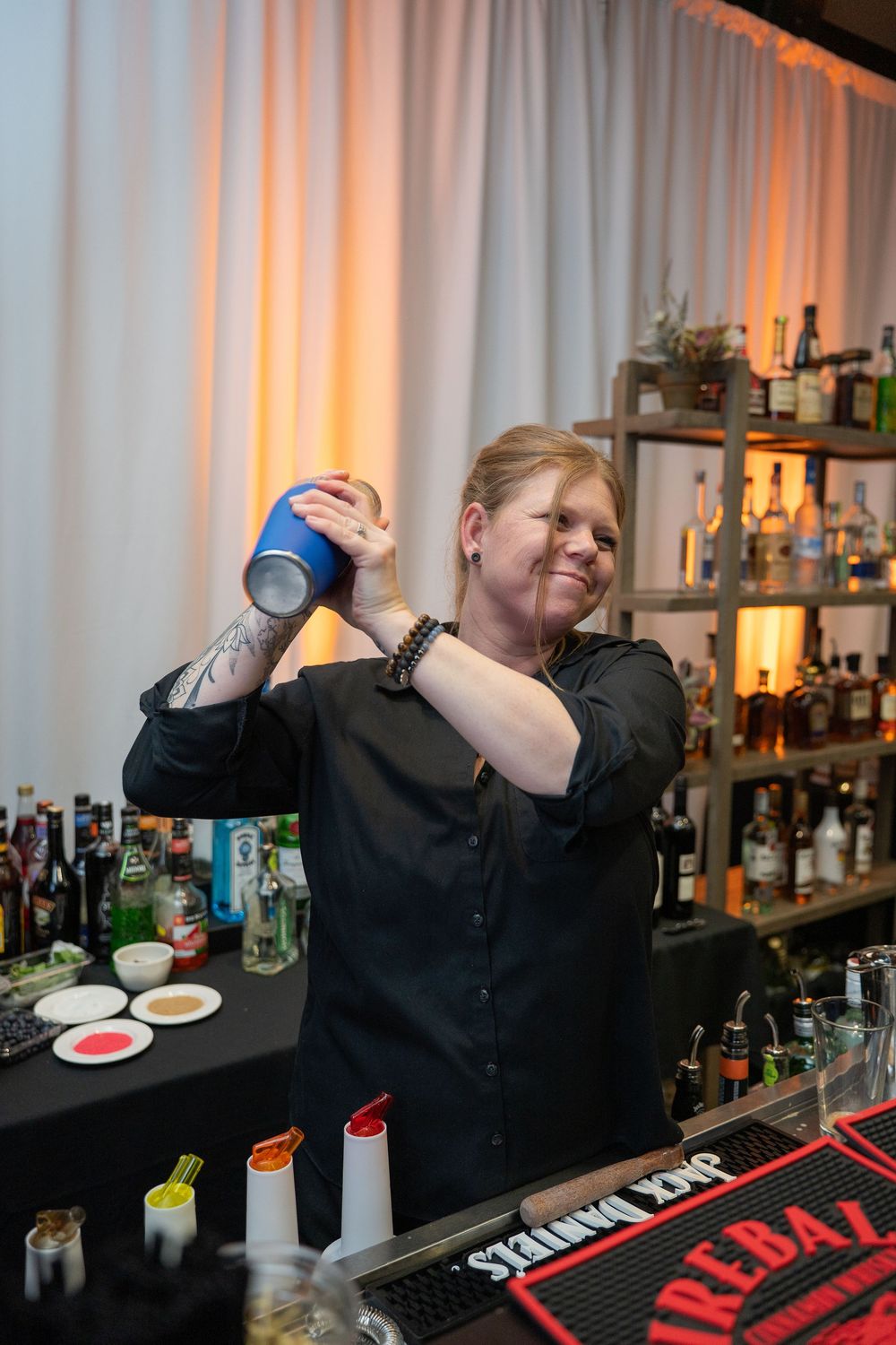 Female bartender shaking a cocktail mixer behind a well-stocked bar at a professional event.