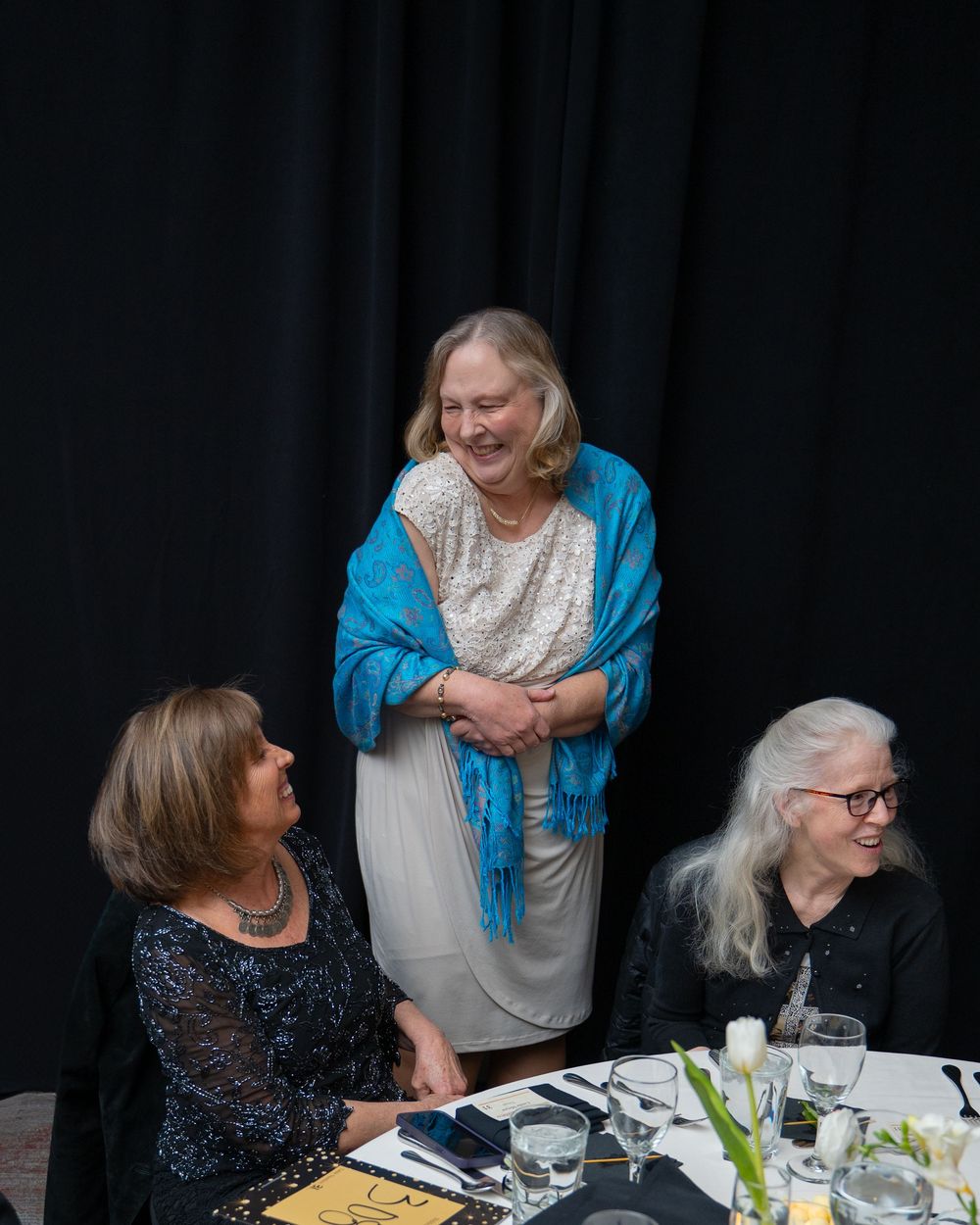 Three older women at a formal dinner event, one standing in a blue shawl, two seated, all smiling warmly.