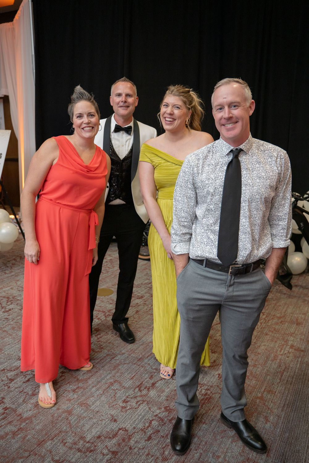 Two couples dressed in formal attire smile together at an elegant event, wearing colorful gowns and suits.