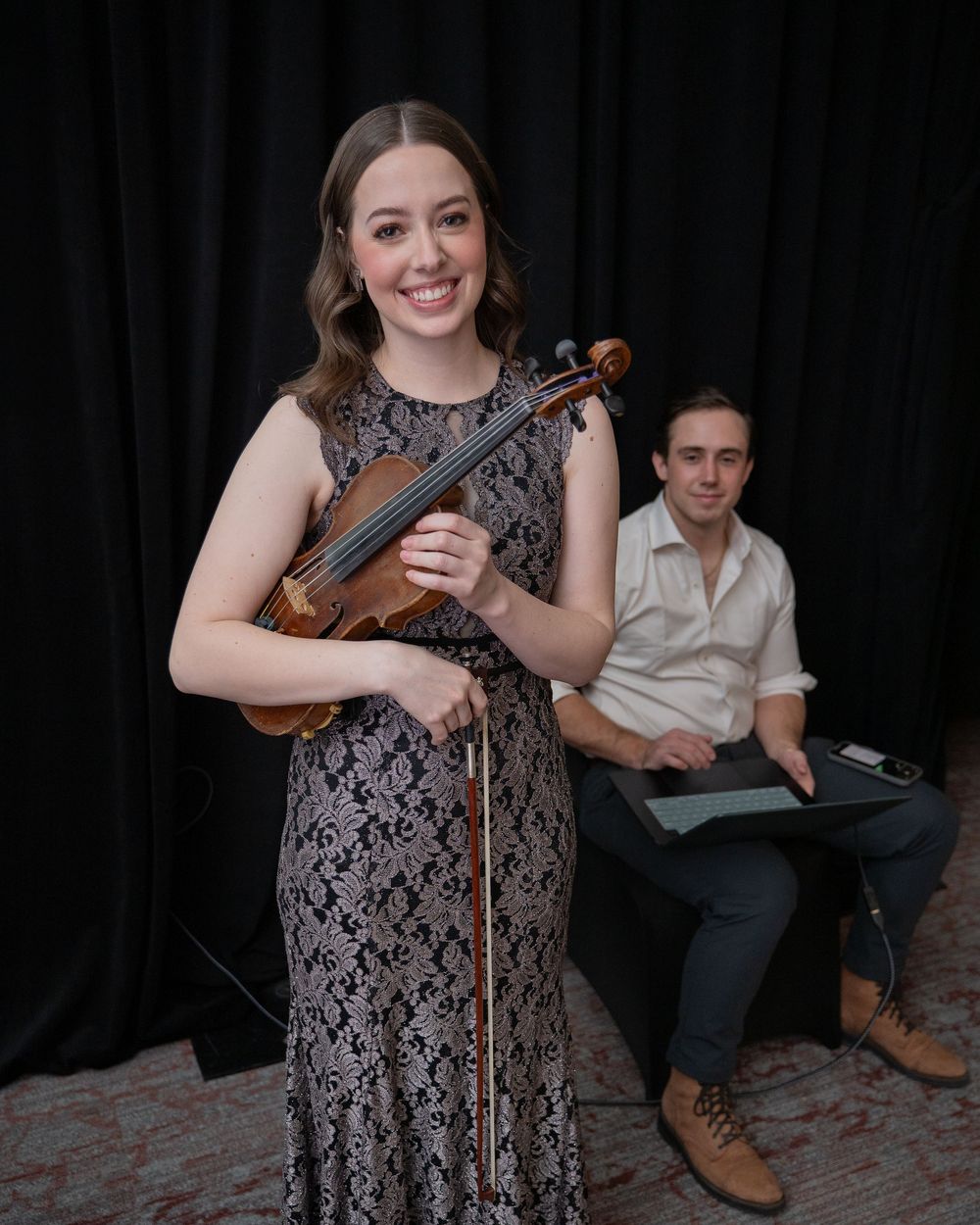 Smiling violinist in lace gown holds violin and bow with male pianist seated in background.