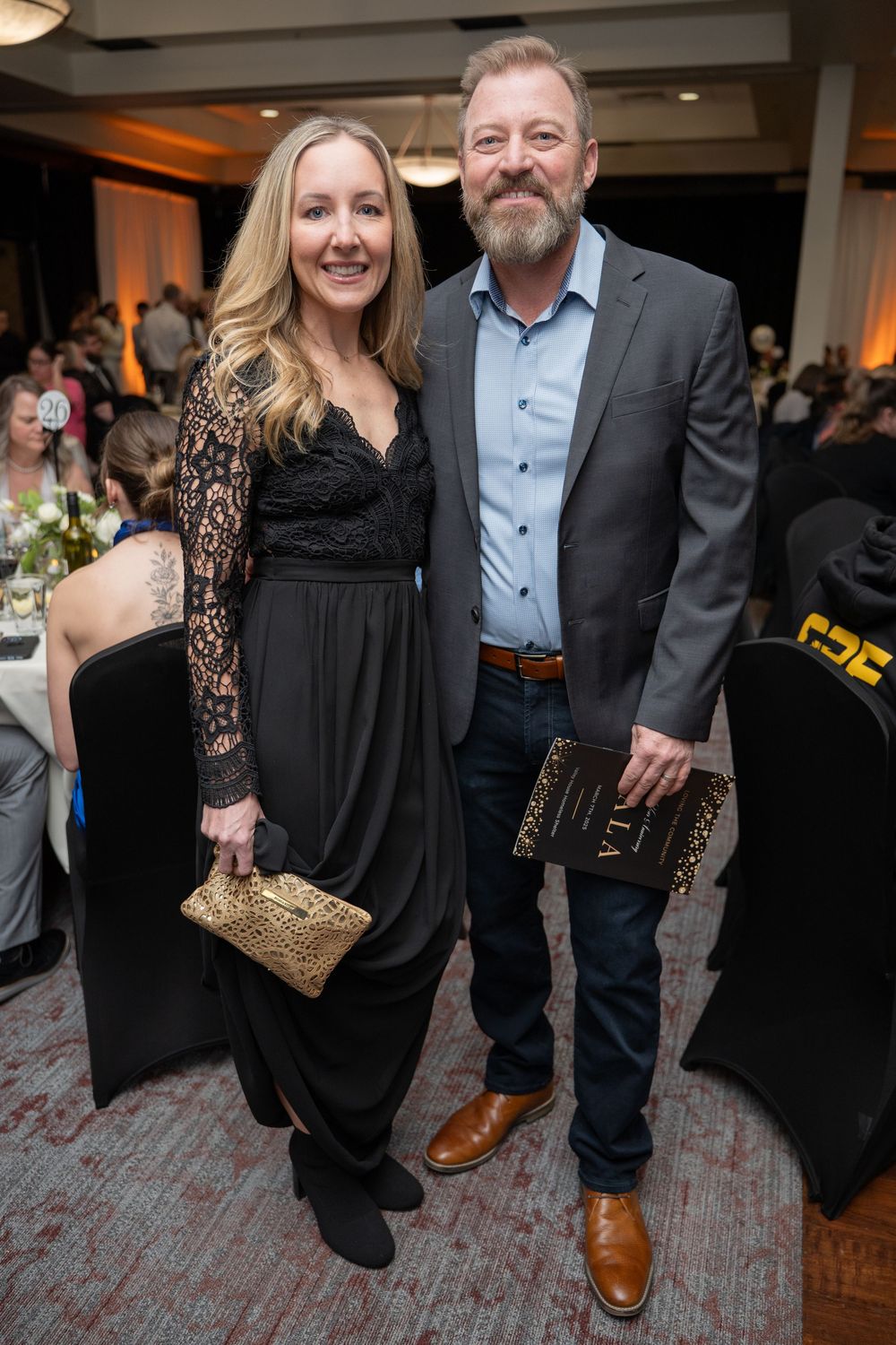 A smiling couple poses together at a formal event, woman in black lace dress with gold clutch, man in gray blazer.