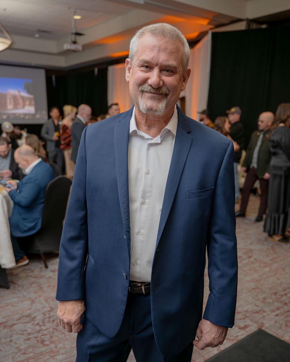Smiling older man with gray hair wearing a navy blue blazer at a formal indoor event.